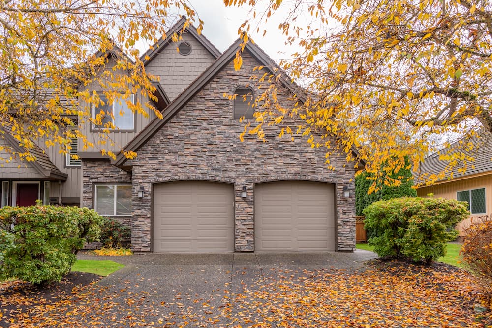 Suburban house with a stone facade and two beige sectional garage doors, emphasizing the importance of regular maintenance and inspecting the door's condition to uphold garage door warranties.